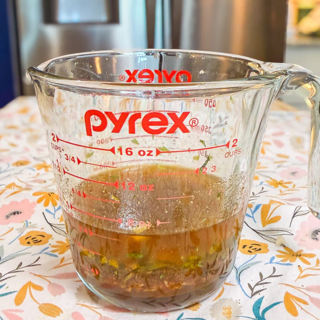 A glass Pyrex measuring cup with brown liquid and herbs inside sits on a floral tablecloth in a kitchen setting. The cup is labeled with red measurement markings up to 2 cups (16 oz).
