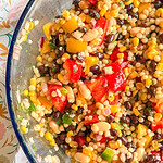 A close-up of a colorful bean and corn salad with diced red and yellow bell peppers, black beans, white beans, pearl couscous, and chopped green onions in a glass bowl.