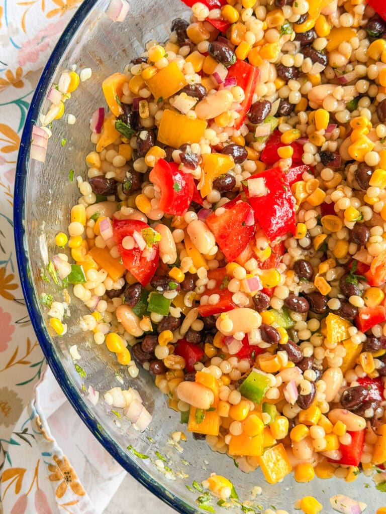 A close-up of a colorful bean and corn salad with diced red and yellow bell peppers, black beans, white beans, pearl couscous, and chopped green onions in a glass bowl.