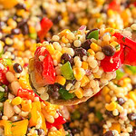 A close-up of a spoonful of colorful bean and vegetable salad, featuring black beans, corn, diced red and green peppers, couscous, and chopped herbs, with more salad blurred in the background.