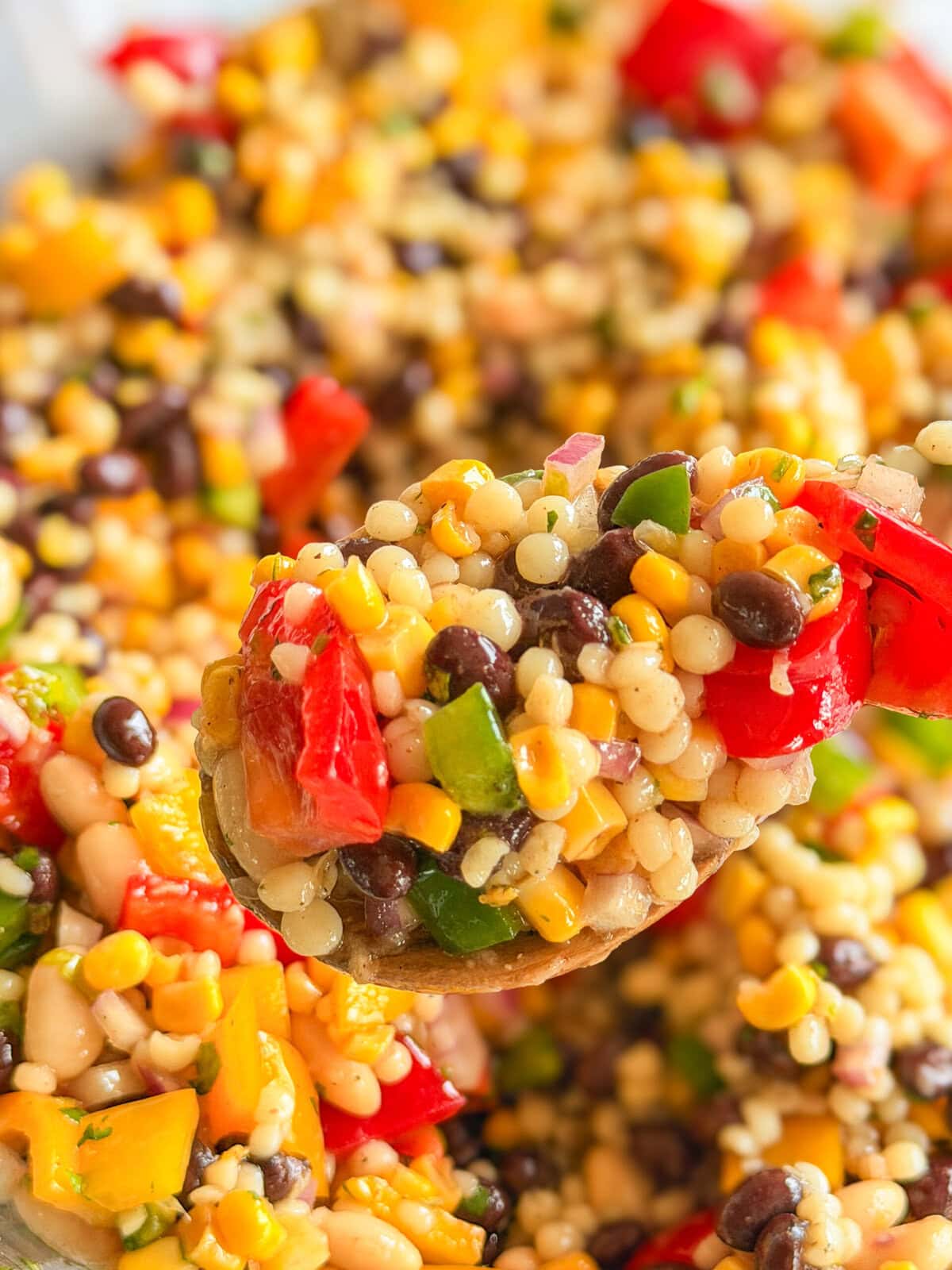 A close-up of a spoonful of colorful bean and vegetable salad, featuring black beans, corn, diced red and green peppers, couscous, and chopped herbs, with more salad blurred in the background.
