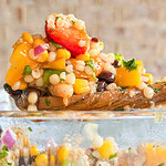 A close-up of a wooden spoon holding a colorful couscous salad with diced tomatoes, yellow bell peppers, red onion, parsley, and black beans, above a glass bowl filled with the same salad.