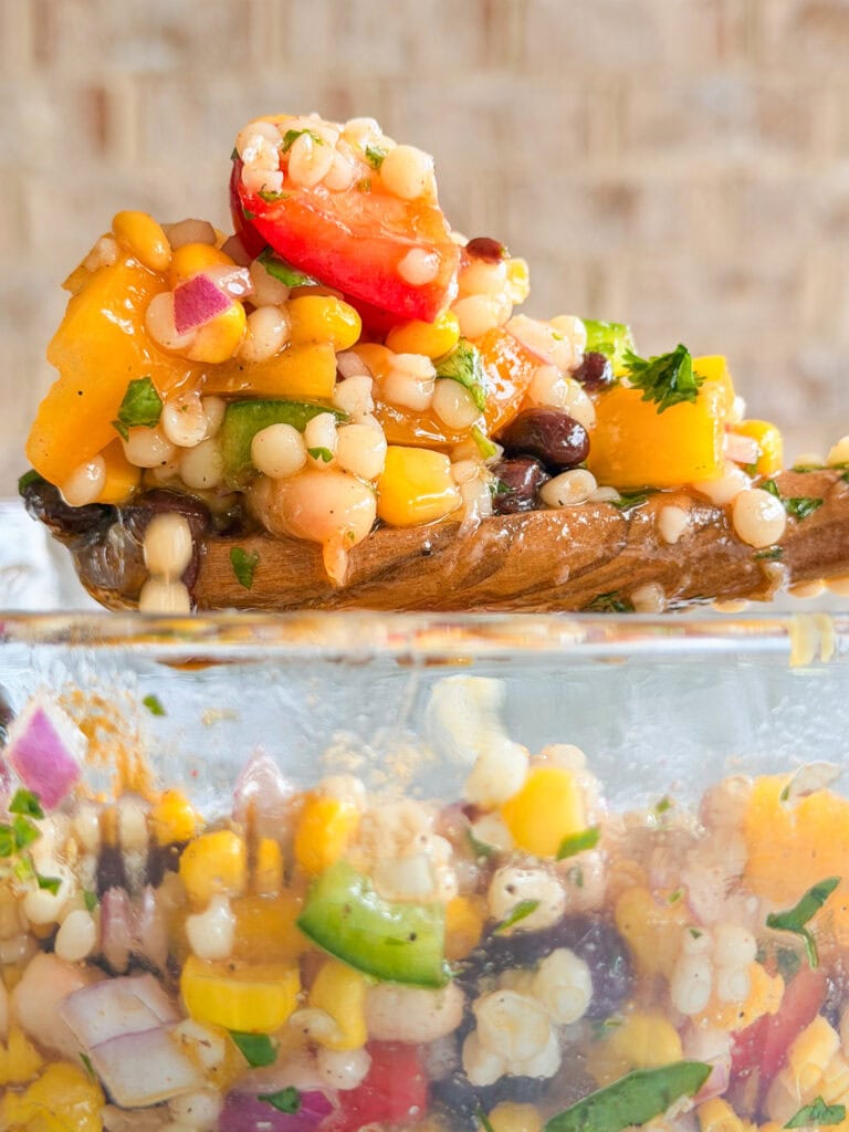A close-up of a wooden spoon holding a colorful couscous salad with diced tomatoes, yellow bell peppers, red onion, parsley, and black beans, above a glass bowl filled with the same salad.
