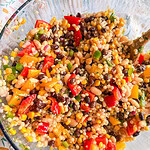 A glass bowl filled with a colorful salad containing black beans, corn, couscous, diced red and yellow bell peppers, chopped green onions, and white beans, all mixed together.