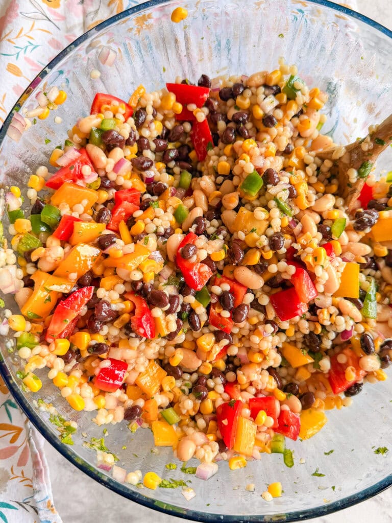 A glass bowl filled with a colorful salad containing black beans, corn, couscous, diced red and yellow bell peppers, chopped green onions, and white beans, all mixed together.