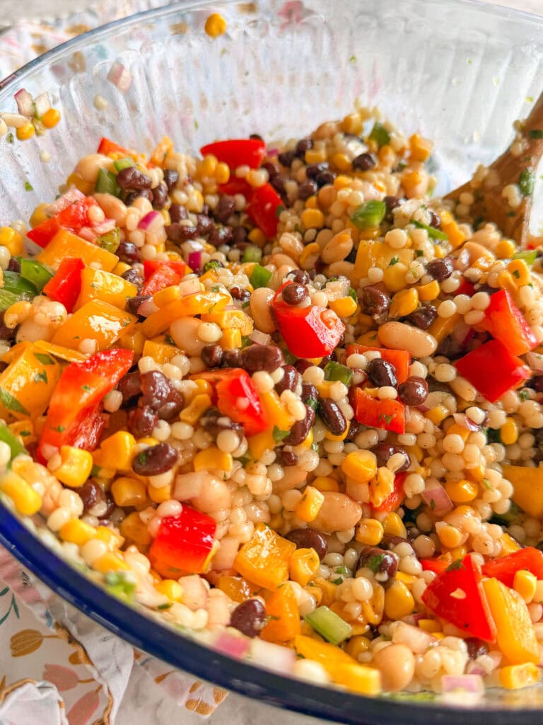 A close-up of a colorful couscous salad with black beans, chickpeas, corn, red bell peppers, diced green onions, and parsley mixed in a clear glass bowl.