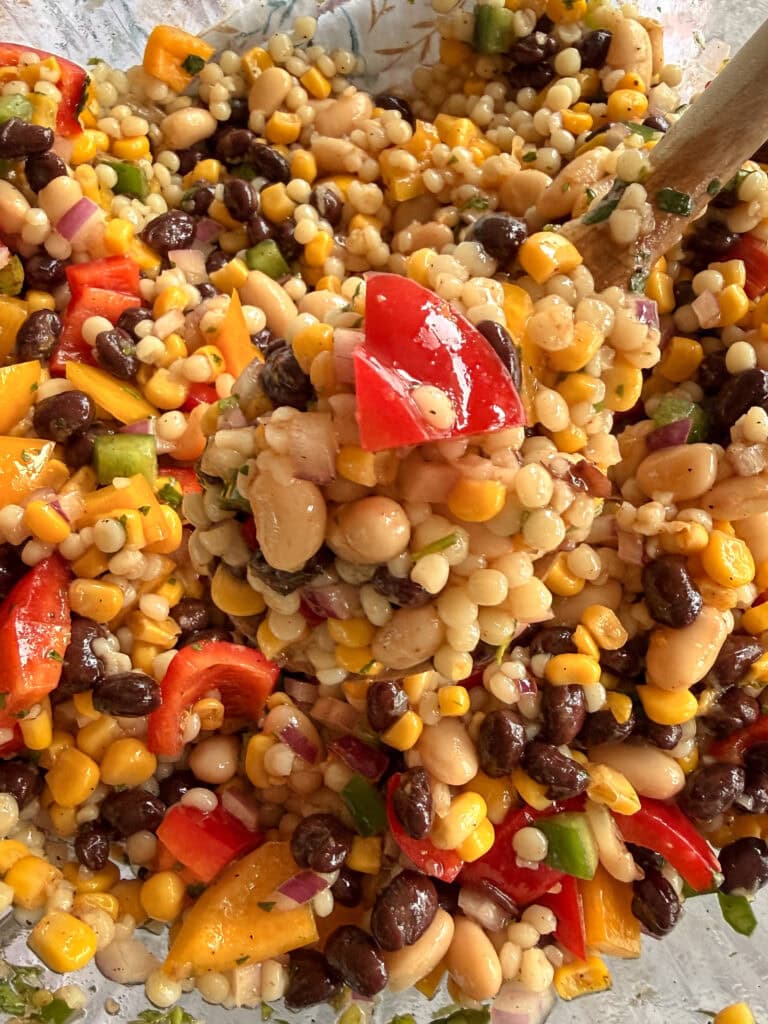 A close-up of a colorful salad with corn, black beans, white beans, chopped red and yellow bell peppers, red onion, and green herbs, being mixed with a wooden spoon.