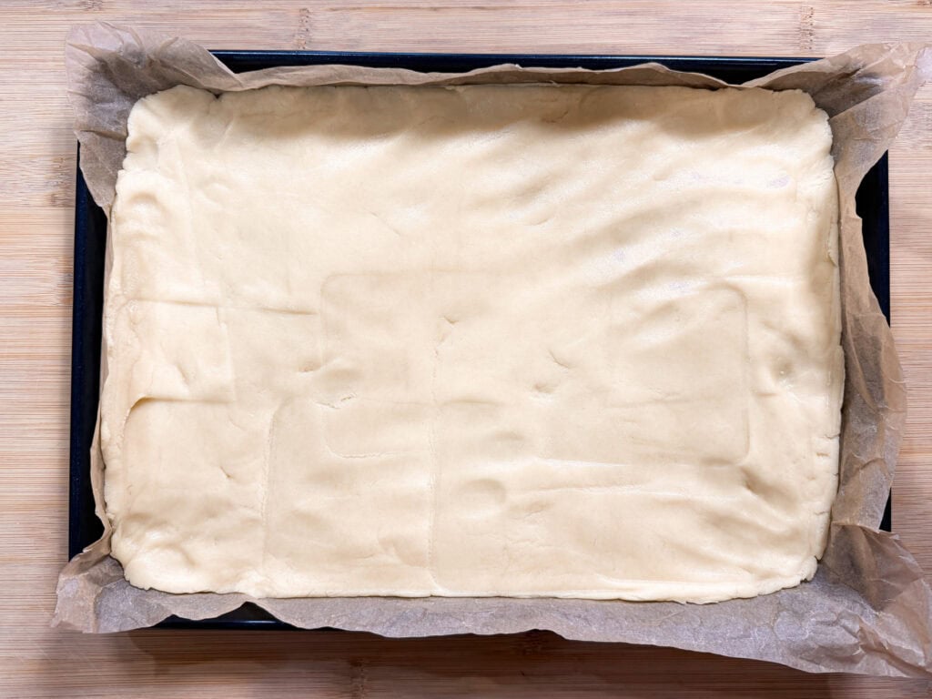 A rectangular baking tray lined with parchment paper and covered with an even layer of raw pastry dough, ready for baking.