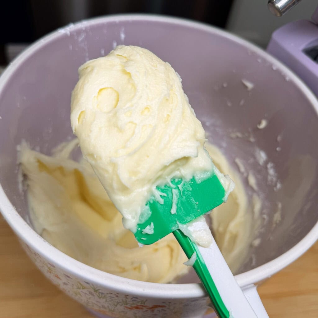A close-up of a green and white spatula holding creamy, whipped frosting above a mixing bowl with more frosting inside. The bowl sits on a light wood surface.