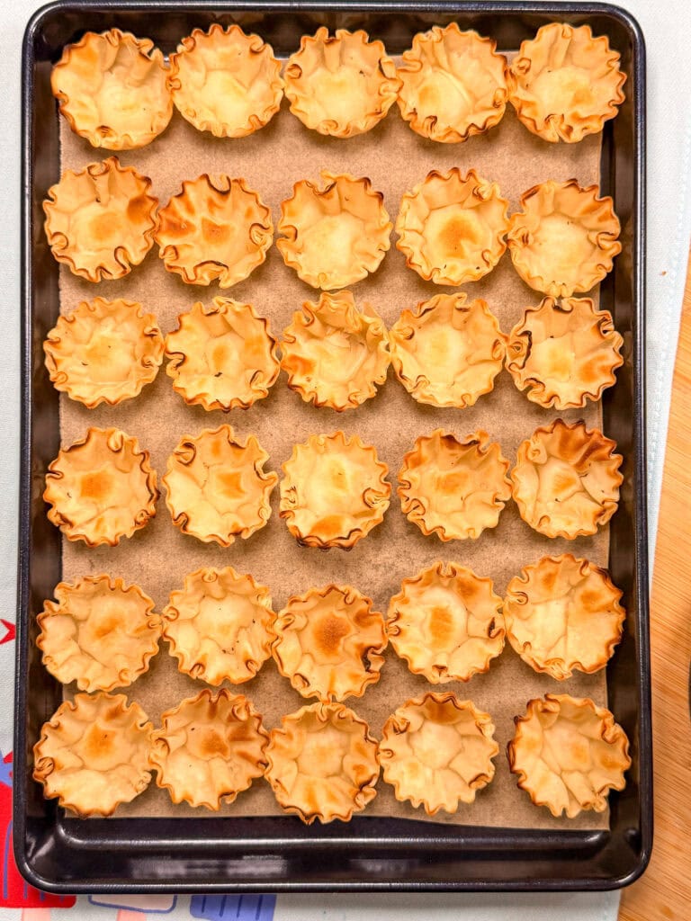 A baking tray lined with parchment paper holds 30 empty, golden-brown baked phyllo pastry cups arranged in neat rows.