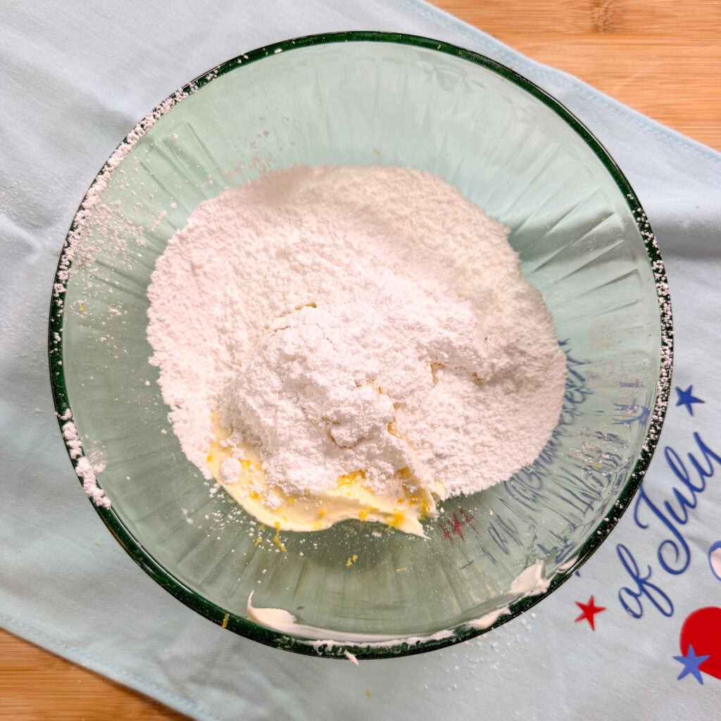 A glass mixing bowl containing powdered sugar and butter sits on a light blue cloth with red and blue stars and of July text, on a wooden surface.