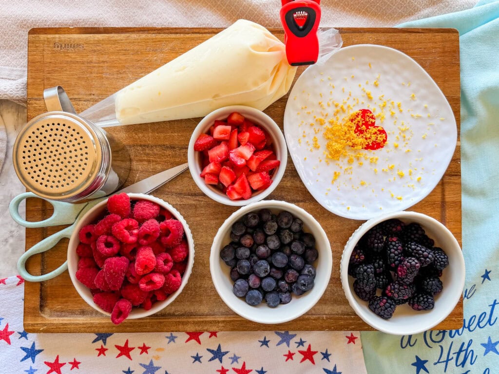 A wooden board with bowls of raspberries, strawberries, blueberries, and blackberries, a piping bag with cream, a shaker, scissors, and a plate with yogurt topped with lemon zest and a heart shape made of strawberries.