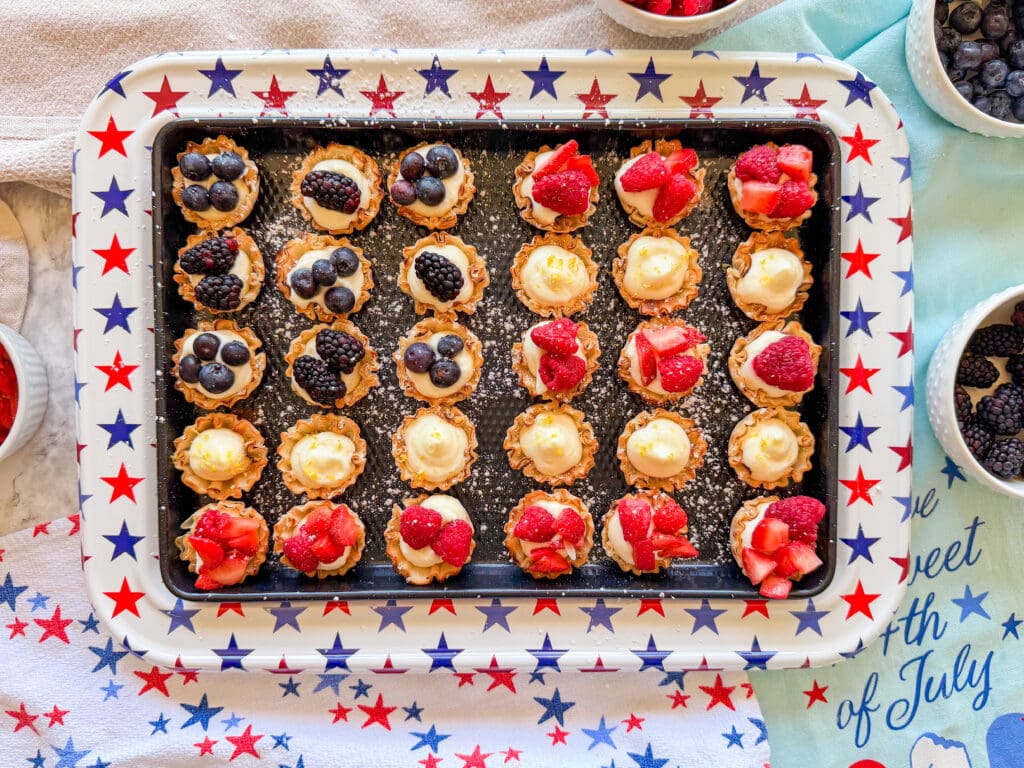 A tray of mini tartlets topped with blueberries, blackberries, and strawberries sits on a star-patterned tray, surrounded by bowls of fresh berries and festive July 4th-themed napkins.