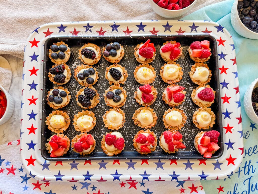 A star-patterned tray holds mini tartlets filled with cream and topped with blueberries, blackberries, and strawberries. Bowls of fresh berries and a blue cloth are nearby.