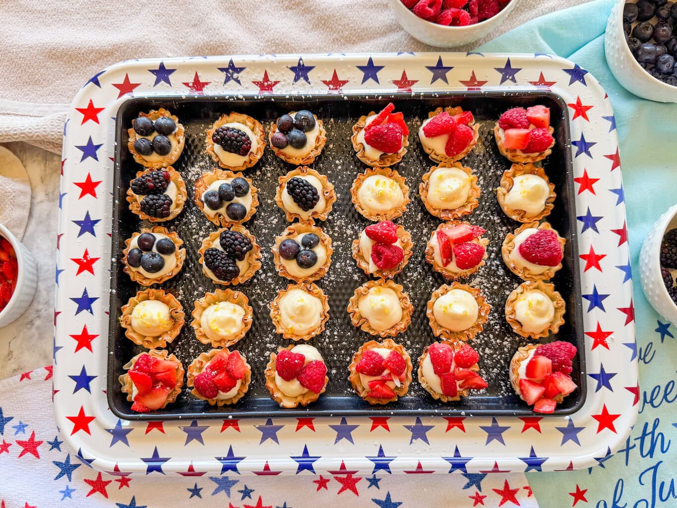 A star-patterned tray holds mini tartlets filled with cream and topped with blueberries, blackberries, and strawberries. Bowls of fresh berries and a blue cloth are nearby.