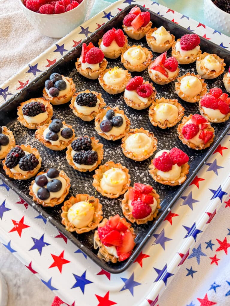 A tray of mini tartlets topped with fresh strawberries, raspberries, blackberries, and blueberries sits on a star-patterned tablecloth, with bowls of berries in the background.