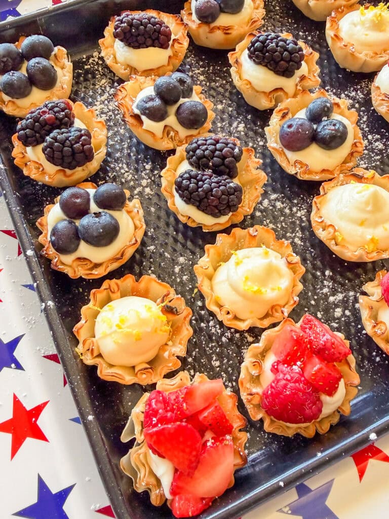 Mini tartlets on a baking tray, topped with fresh berries like strawberries, blueberries, and blackberries over cream filling. Some tarts are garnished with lemon zest. The tray is on a star-patterned tablecloth.