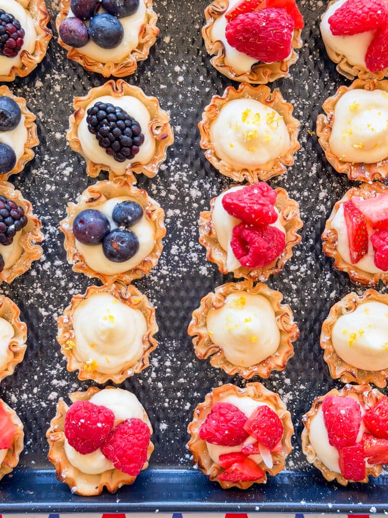 Mini tart shells filled with cream and topped with assorted berries, including strawberries, raspberries, blueberries, and blackberries, arranged neatly on a tray dusted with powdered sugar.