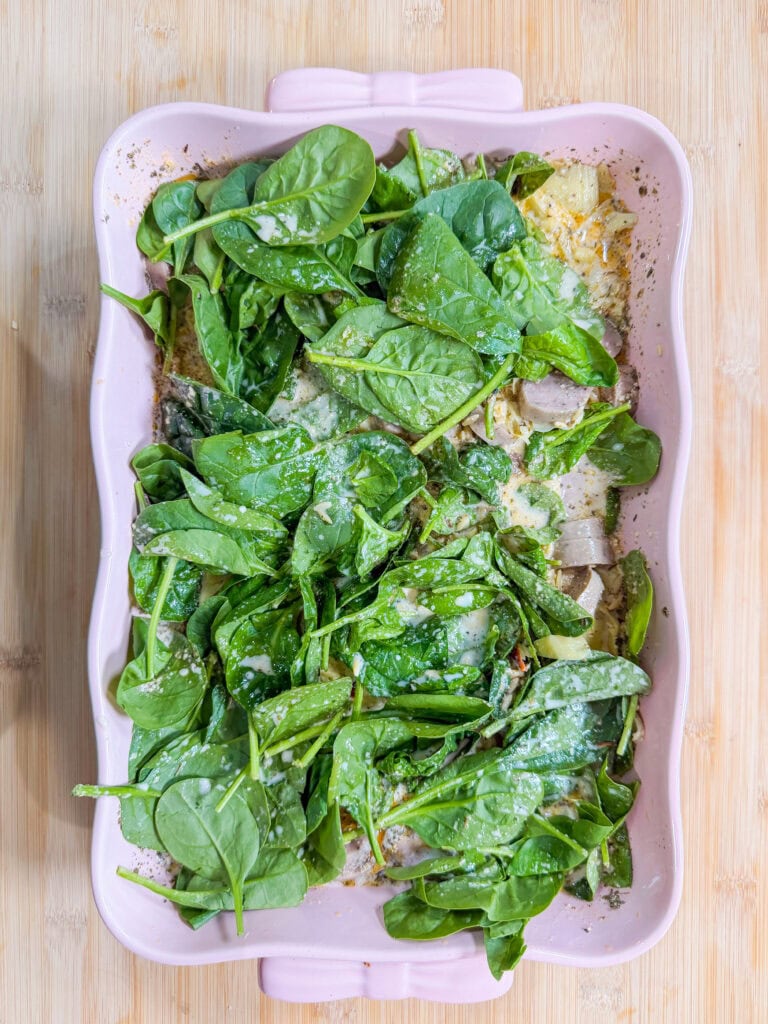A pink baking dish filled with fresh spinach leaves, sliced onions, shredded cheese, and a creamy sauce, all layered and ready to be cooked, sits on a light wooden surface.