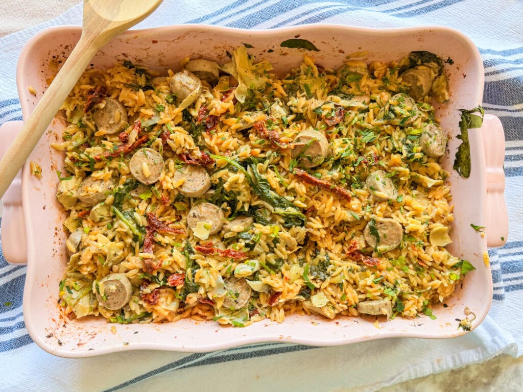 A casserole dish filled with baked orzo pasta, sliced sausage, spinach, sun-dried tomatoes, and herbs, placed on a striped cloth with a wooden spoon resting on the side.