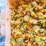 A pink baking dish filled with cooked orzo pasta, sliced sausages, sun-dried tomatoes, and leafy greens, set on a blue and white striped cloth.
