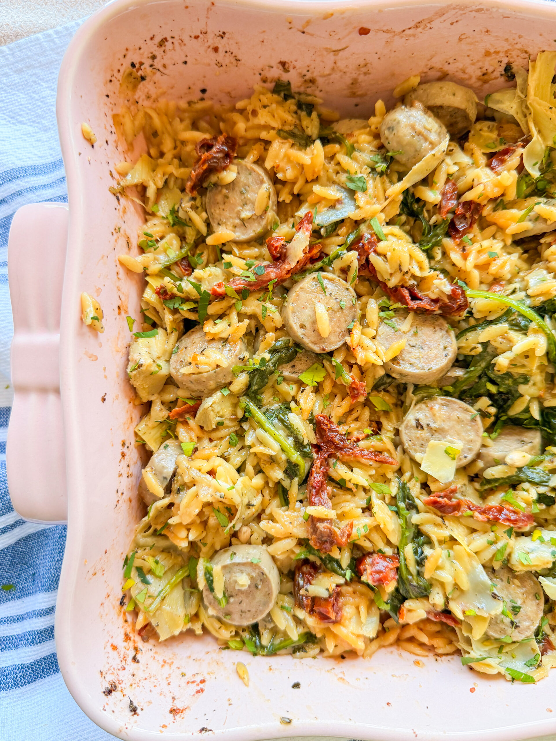 A pink baking dish filled with cooked orzo pasta, sliced sausages, sun-dried tomatoes, and leafy greens, set on a blue and white striped cloth.