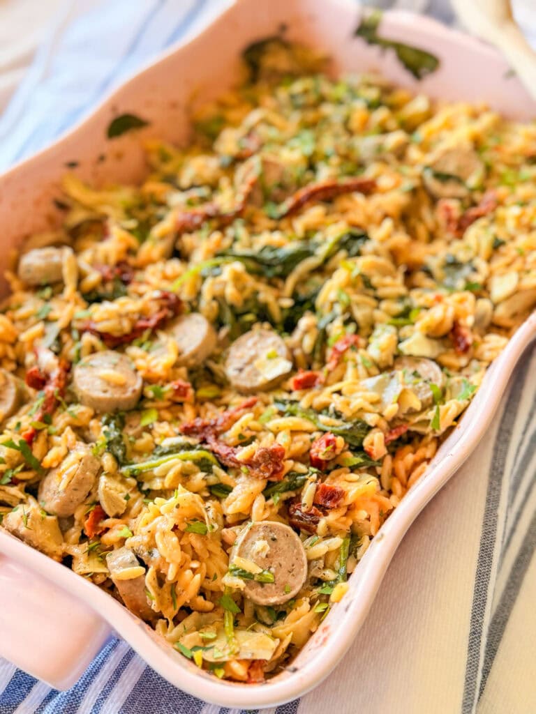 A baked casserole dish filled with orzo pasta, sliced sausage, sun-dried tomatoes, spinach, and zucchini, set on a striped cloth.