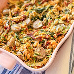 A casserole dish filled with orzo pasta, sliced sausage, spinach, sun-dried tomatoes, and herbs, arranged on a striped cloth.