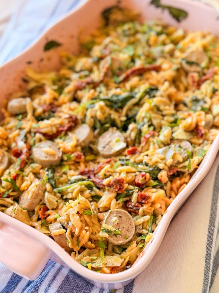A casserole dish filled with orzo pasta, sliced sausage, spinach, sun-dried tomatoes, and herbs, arranged on a striped cloth.