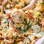 A close-up of a wooden spoon lifting creamy orzo pasta mixed with spinach, sun-dried tomatoes, and sliced sausage from a dish.