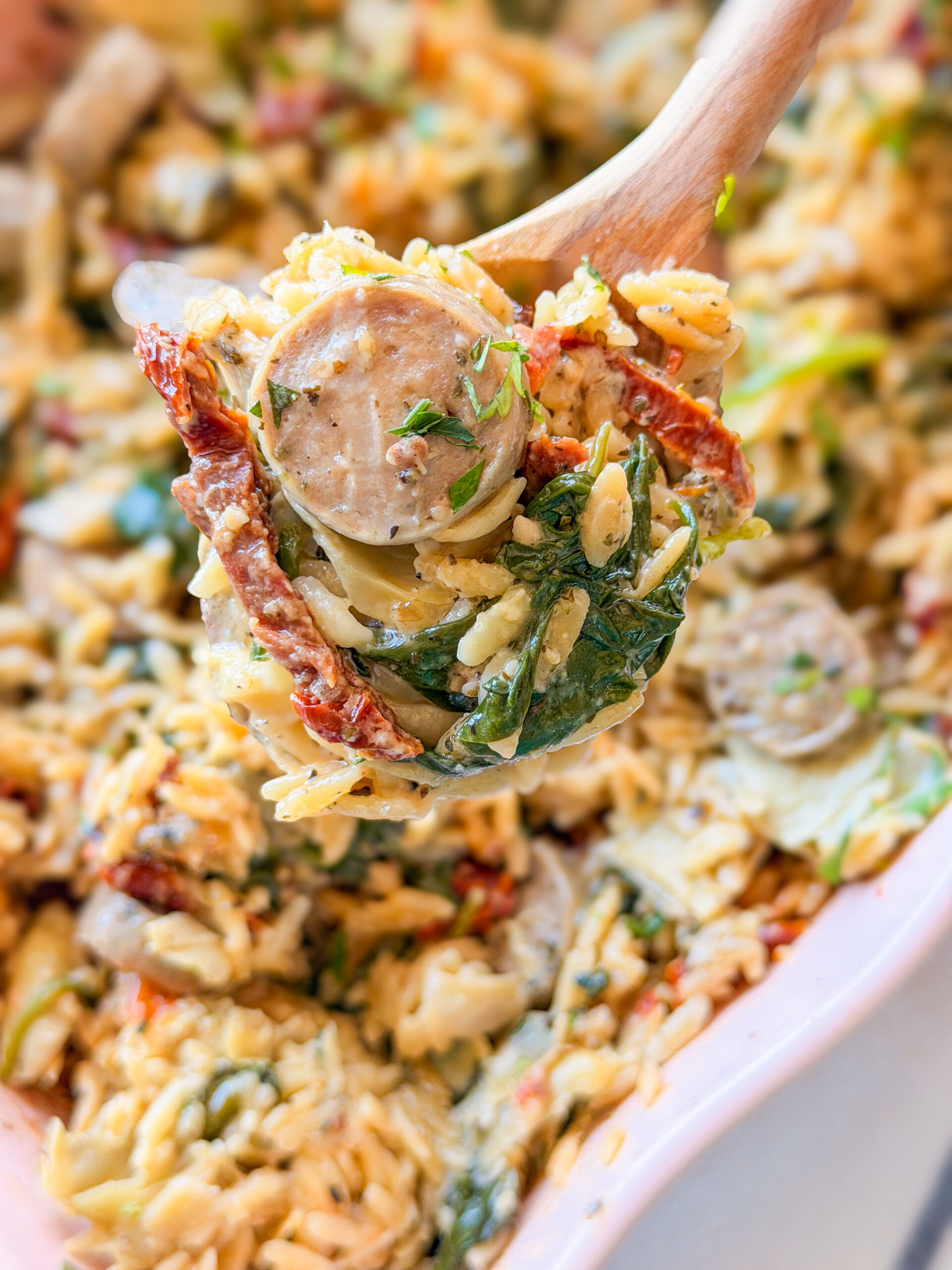 A close-up of a wooden spoon lifting creamy orzo pasta mixed with spinach, sun-dried tomatoes, and sliced sausage from a dish.