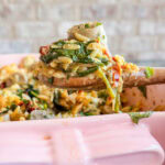 A wooden spoon holds a serving of baked orzo pasta with sausage, spinach, and sun-dried tomatoes above a pink baking dish. The dish appears cheesy and garnished with fresh herbs.