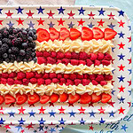 A rectangular tray decorated with red and blue stars displays a dessert arranged as the American flag using blackberries, strawberries, raspberries, and piped white cream.