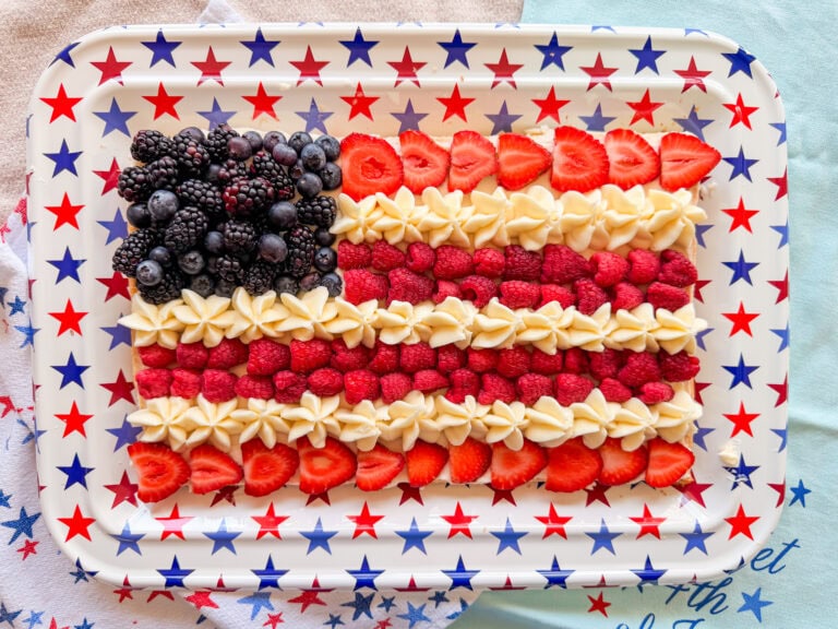 A rectangular tray decorated with red and blue stars displays a dessert arranged as the American flag using blackberries, strawberries, raspberries, and piped white cream.