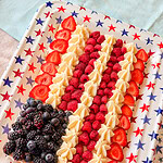 A rectangular dessert on a star-patterned tray decorated like an American flag, with blackberries and blueberries in one corner, and rows of raspberries, strawberries, and piped cream forming stripes.