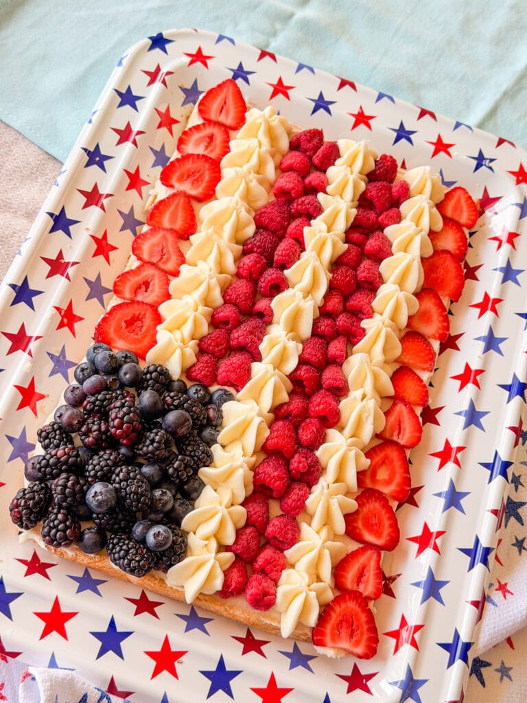 A rectangular dessert on a star-patterned tray decorated like an American flag, with blackberries and blueberries in one corner, and rows of raspberries, strawberries, and piped cream forming stripes.