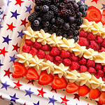 A rectangular dessert decorated like an American flag with blackberries and blueberries for the top left, raspberries and sliced strawberries for red stripes, and white icing for white stripes, on a star-patterned tray.