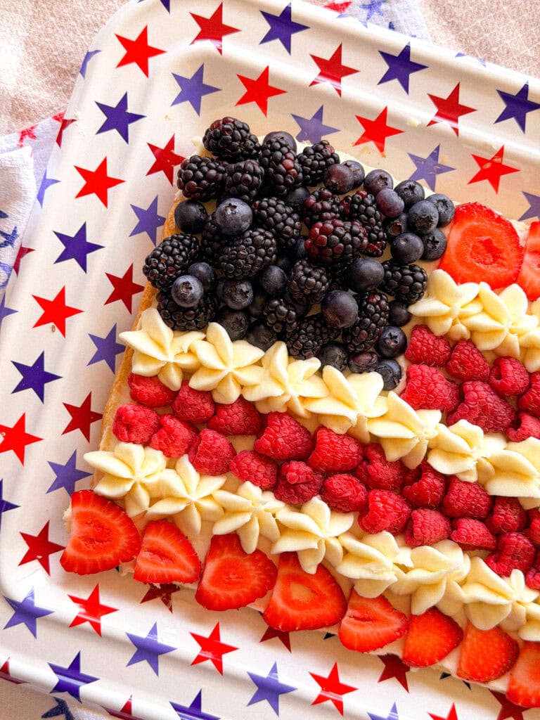 A rectangular dessert decorated like an American flag with blackberries and blueberries for the top left, raspberries and sliced strawberries for red stripes, and white icing for white stripes, on a star-patterned tray.