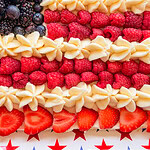 A dessert decorated like the American flag, with blueberries and blackberries in the top left corner, strawberries and raspberries as red stripes, and piped white cream as white stripes. The dessert is on a star-patterned tray.