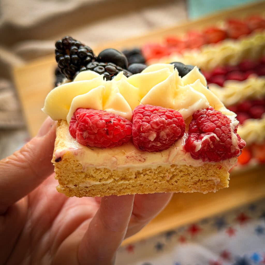 A hand holds a square piece of dessert with a golden crust, creamy topping, fresh raspberries, blueberries, blackberries, and piped whipped cream, with a tray of similar treats blurred in the background.