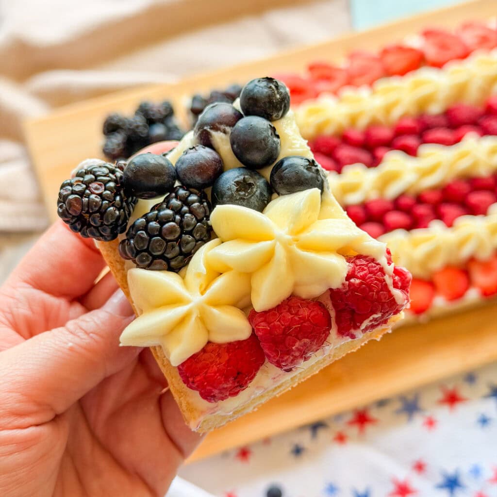 A hand holds a dessert decorated as an American flag with blueberries, blackberries, raspberries, and white frosting stars. The dessert sits on a wooden board with more pieces in the background.
