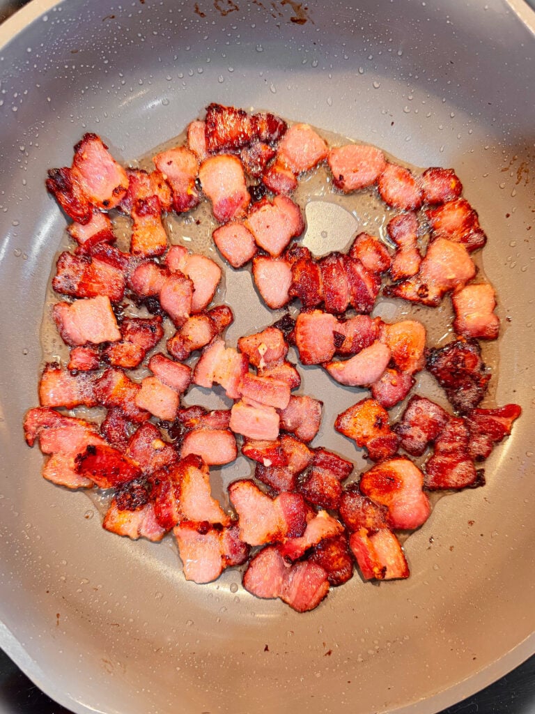 Crispy bacon pieces cooking in a frying pan, arranged in a circular pattern with some oil droplets visible on the pan’s surface.