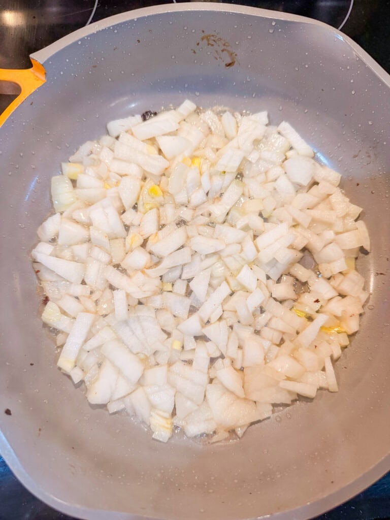 Chopped onions are sautéing in a gray frying pan on a stovetop, starting to turn translucent as they cook in hot oil.