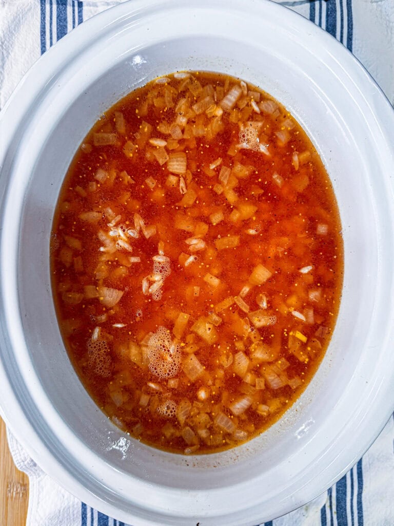 A white slow cooker filled with a soup or broth containing chopped onions and a reddish-orange liquid, placed on a striped cloth.