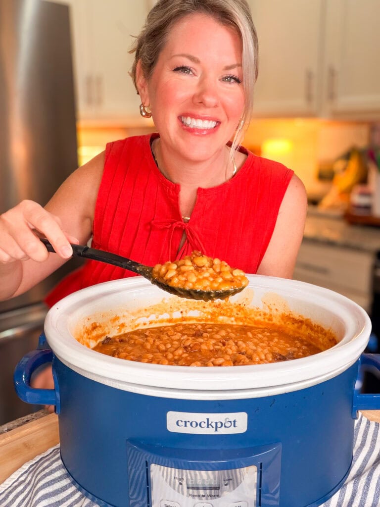 A smiling woman in a red sleeveless top holds a spoonful of baked beans over a blue Crockpot filled with beans in a kitchen setting.