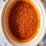 A white slow cooker filled with baked beans in a rich, reddish-brown sauce sits on a striped blue and white cloth, with a green napkin and wooden spoon nearby.