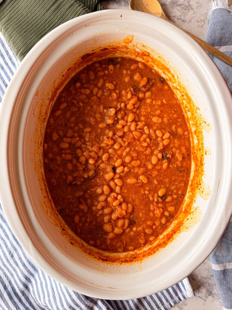 A white slow cooker filled with baked beans in a rich, reddish-brown sauce sits on a striped blue and white cloth, with a green napkin and wooden spoon nearby.