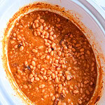 A close-up of baked beans in a white slow cooker, showing a thick, reddish sauce and tender beans. Some pieces of seasoning or meat are visible among the beans.