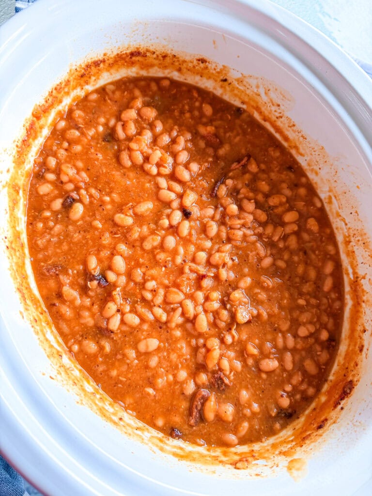 A close-up of baked beans in a white slow cooker, showing a thick, reddish sauce and tender beans. Some pieces of seasoning or meat are visible among the beans.