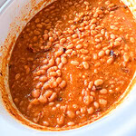 A close-up of baked beans in a white slow cooker, showing a thick, orange-brown sauce with visible beans and bits of meat.